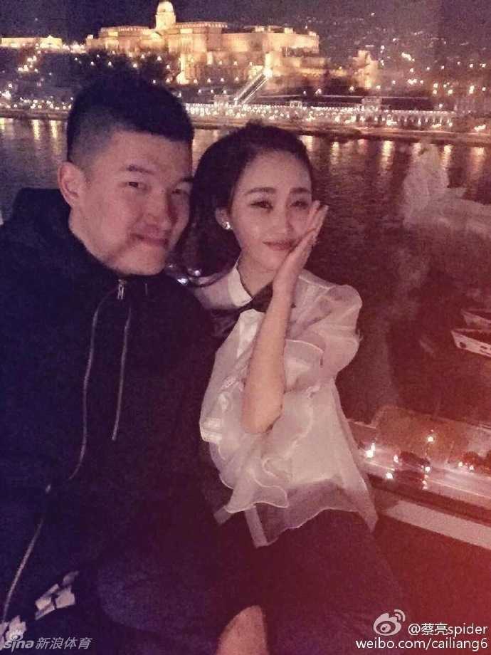 两人在一个充满设计感的卡通雕塑旁合影，蔡亮搞怪地模仿着雕塑的动作，逗得身边的未婚妻开怀大笑，画面充满了青春的活力与俏皮。